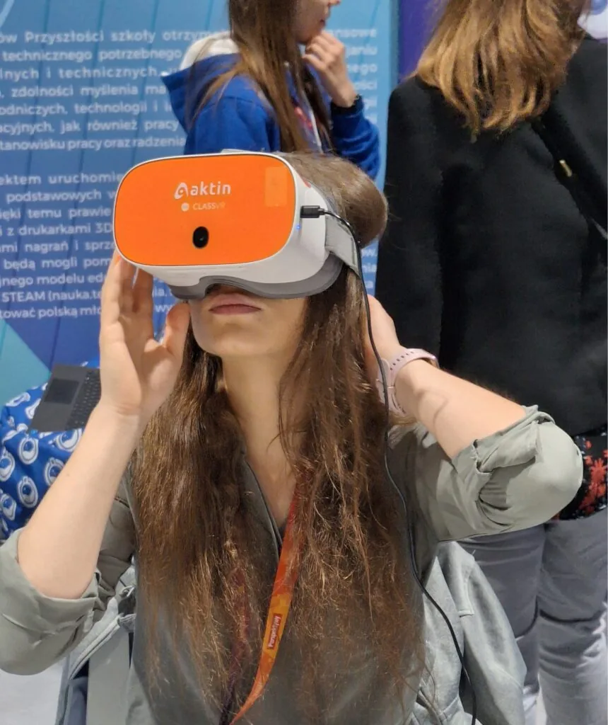 Uczestniczka konferencji przymierza gogle VR na stoisku wystawowym; w tle plansza o wyposażeniu szkół w sprzęt techniczny