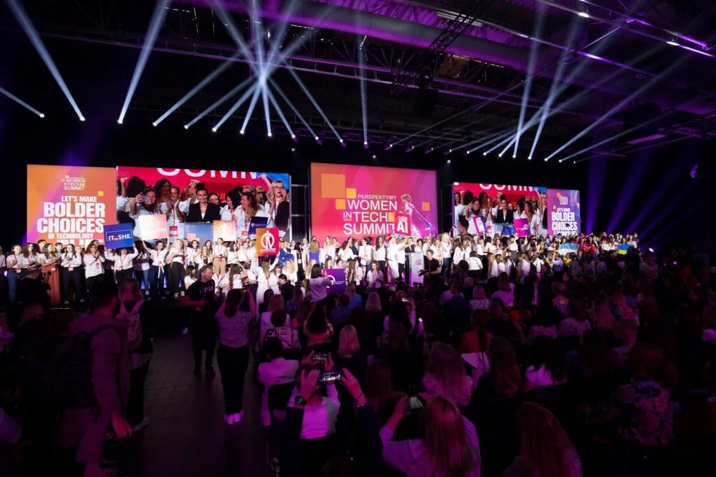 Otwarcie konferencji Perspektywy Women in Tech Summit: pełna hala, snopy światła nad publicznością i rząd osób na scenie przed ekranami z hasłem „Let's make bolder choices in technology”