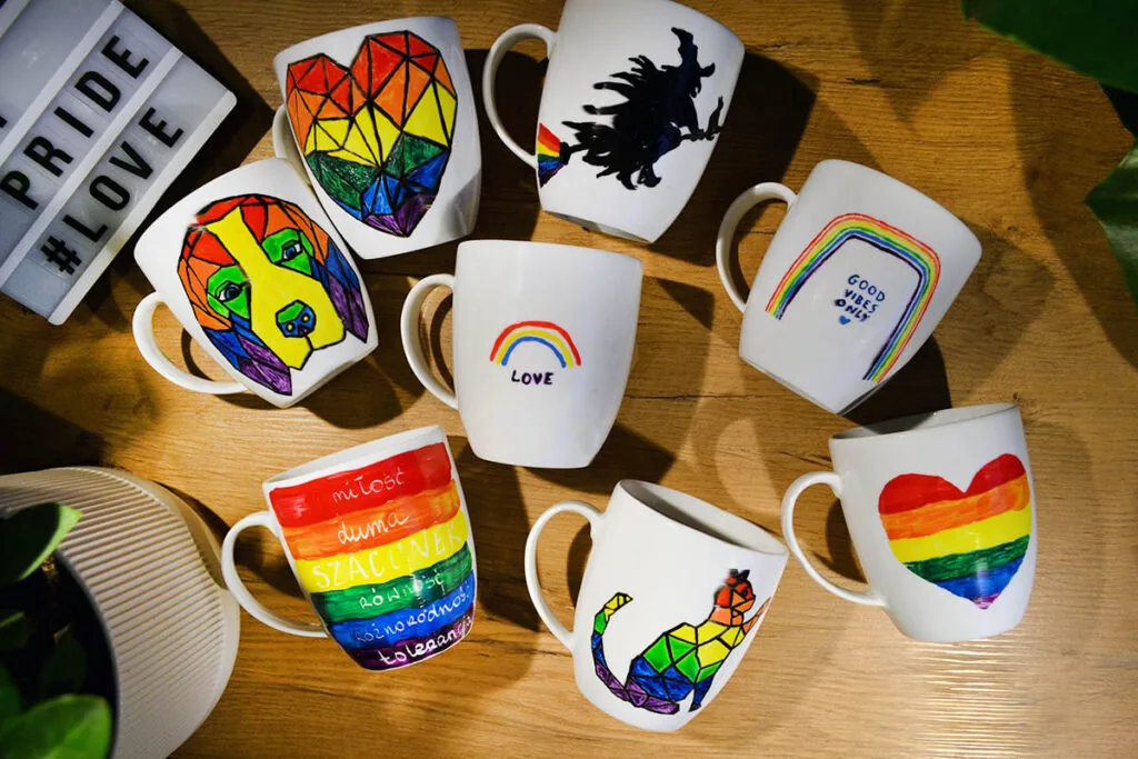 Osiem białych kubków na drewnianym blacie, każdy pomalowany w tęczowe wzory: serca, pies, kot, napisy „love”, „good vibes only” oraz „miłość, duma, szacunek, równość, różnorodność, tolerancja”. Obok tablica z napisem „#PRIDE #LOVE”